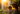 A warmly lit close-up of a smiling mother in a straw hat holding her baby outdoors during golden hour. The intense sunlight backlights them, creating a radiant halo effect around their hair and shoulders, with shimmering water droplets visible against lush green foliage in the left background. The baby, dressed in light fabric, looks towards the viewer with wide eyes while the mother, wearing a floral-patterned dress, gazes lovingly at the child.