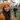 A young woman with long dark hair, wearing an orange knit beanie and sweater, smiles brightly at the camera in the foreground. She is surrounded by a large pile of brown and orange autumn leaves on the ground. In the blurred background, a man is seen raking leaves in front of a house, suggesting a fall yard work scene.