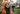 A young woman with long dark hair, wearing an orange knit beanie and sweater, smiles brightly at the camera in the foreground. She is surrounded by a large pile of brown and orange autumn leaves on the ground. In the blurred background, a man is seen raking leaves in front of a house, suggesting a fall yard work scene.