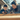 A sun-kissed close-up of a mother and her young son collaboratively working on a woodworking project outdoors. They are both looking down, focused on a light wooden plank, each holding a part of an orange and black measuring tool. The mother wears a grey top with her dark hair tied up, and the boy wears a dark blue long-sleeved shirt, both illuminated by warm light.