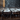 A close-up view captures numerous white water droplets falling rapidly and splashing onto a dark brown wooden surface. The wet planks show reflections, small bubbles, and water dripping off the edges, highlighting the texture and dynamic interaction of water and wood.
