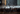 A close-up view captures numerous white water droplets falling rapidly and splashing onto a dark brown wooden surface. The wet planks show reflections, small bubbles, and water dripping off the edges, highlighting the texture and dynamic interaction of water and wood.