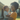 A joyful African American mother with an afro hairstyle, wearing an orange shirt, holds her baby outdoors in warm, golden light. Both mother and baby are smiling broadly and looking into each other's eyes, conveying deep affection. The background is softly blurred, showing parts of a light-colored house and green foliage, indicating a domestic or suburban setting.