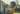 A joyful African American mother with an afro hairstyle, wearing an orange shirt, holds her baby outdoors in warm, golden light. Both mother and baby are smiling broadly and looking into each other's eyes, conveying deep affection. The background is softly blurred, showing parts of a light-colored house and green foliage, indicating a domestic or suburban setting.