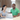 A happy diverse couple, an Asian woman and a Black man, are sitting at a white kitchen island, looking at a silver MacBook laptop with bright smiles. The modern kitchen features dark wood cabinets, stainless steel appliances, and sleek light fixtures, illuminated by bright, clear lighting. The woman wears a grey t-shirt and the man a green polo shirt with a small logo, both appearing engaged and joyful.