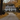 A stately white colonial-style house with dark shutters stands at the end of a long, curving gravel driveway, surrounded by a vibrant autumn landscape. The ground and trees are abundant with golden and yellow fallen leaves, and a dark-colored dog sits on the grass beside the driveway, facing away from the viewer. The symmetrical house features multiple windows and a prominent central entrance, all set under a soft, overcast sky.