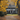 A picturesque two-story white house with black shutters and a front portico is framed by towering trees adorned with vibrant golden-yellow autumn leaves. A winding dirt path, covered in fallen brown leaves, leads to a green lawn where two dogs, one black and white and one black, rest peacefully in a patch of sunlight. The warm, soft lighting highlights the rich fall colors and creates an inviting, serene atmosphere.