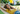 A young woman with long, wavy brown hair lies peacefully in a light-colored woven hammock, with her eyes closed and a gentle smile. She is wearing a yellow t-shirt and blue jeans, illuminated by warm sunlight. The background is a softly blurred outdoor garden featuring lush green foliage and a bright yellow palm frond, creating a serene summer atmosphere.