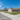 A wide-angle view of a serene suburban street under a bright blue sky with scattered white clouds. A row of single-story houses with light-colored walls, some with brick accents, and brown tiled roofs line the right side of the street, each fronted by a neatly mowed green lawn and a concrete sidewalk. The paved street stretches into the distance on the left, with shadows cast across it.