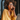 A close-up vertical shot of a young Black woman with voluminous natural curly hair and round tortoiseshell glasses, smiling serenely with her eyes closed in warm golden sunlight. She holds a silver smartphone to her right ear, appearing to be enjoying a pleasant phone conversation. She wears a mustard yellow button-down shirt, and a silver laptop is partially visible on a light wooden table in the bottom right.