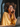 A close-up vertical shot of a young Black woman with voluminous natural curly hair and round tortoiseshell glasses, smiling serenely with her eyes closed in warm golden sunlight. She holds a silver smartphone to her right ear, appearing to be enjoying a pleasant phone conversation. She wears a mustard yellow button-down shirt, and a silver laptop is partially visible on a light wooden table in the bottom right.