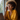 A close-up of a smiling Black woman with prominent curly hair and round glasses, holding a dark-colored smartphone to her right ear. She is wearing a mustard yellow button-down shirt and delicate gold necklaces, illuminated by natural light from a window to her left. A blurred silver laptop and wooden desk are visible in the foreground, suggesting a professional yet comfortable remote work environment.