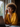 A close-up of a smiling Black woman with prominent curly hair and round glasses, holding a dark-colored smartphone to her right ear. She is wearing a mustard yellow button-down shirt and delicate gold necklaces, illuminated by natural light from a window to her left. A blurred silver laptop and wooden desk are visible in the foreground, suggesting a professional yet comfortable remote work environment.