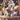 A close-up vertical shot of a brown wicker basket filled with decorative Easter items. Inside, several pastel and speckled eggs are arranged around two small ceramic houses, one cream-yellow with a brown roof and one white with a brown roof, all resting on green faux moss. In the soft-focus background, spring branches with tiny yellow and white blossoms are visible, set against a dark wooden surface.