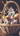 A close-up vertical shot of a brown wicker basket filled with decorative Easter items. Inside, several pastel and speckled eggs are arranged around two small ceramic houses, one cream-yellow with a brown roof and one white with a brown roof, all resting on green faux moss. In the soft-focus background, spring branches with tiny yellow and white blossoms are visible, set against a dark wooden surface.