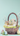 A charming brown wicker basket is centered against a soft light blue background, filled with two miniature pastel-colored houses. Artificial green grass and several speckled Easter eggs in soft hues of blue, green, and pink are nestled around the houses within the basket. Pussy willow branches emerge from behind the houses, and additional pastel Easter eggs are scattered around the base of the basket on the matching light blue surface, creating a whimsical spring holiday scene.
