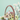 A vertical image featuring a small, yellow and brown miniature house model placed inside a brown wicker basket. The basket contains vibrant green moss, artificial grass, and several pastel Easter eggs in shades of light blue, yellow, and speckled white. A branch with pale pink and white flowers extends from the right side of the basket, all set against a solid, soft mint green background.