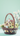 A vertical image featuring a small, yellow and brown miniature house model placed inside a brown wicker basket. The basket contains vibrant green moss, artificial grass, and several pastel Easter eggs in shades of light blue, yellow, and speckled white. A branch with pale pink and white flowers extends from the right side of the basket, all set against a solid, soft mint green background.