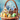 A detailed close-up of a woven wicker basket filled with Easter decorations. Inside the basket, three miniature, colorful houses are nestled in vibrant green moss, surrounded by various brightly decorated Easter eggs. The basket sits on a rustic wooden surface with more decorated eggs scattered nearby, all set against a soft, painterly blue and green background.