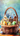A detailed close-up of a woven wicker basket filled with Easter decorations. Inside the basket, three miniature, colorful houses are nestled in vibrant green moss, surrounded by various brightly decorated Easter eggs. The basket sits on a rustic wooden surface with more decorated eggs scattered nearby, all set against a soft, painterly blue and green background.