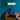 A young Black man with a beard and short, styled hair sits relaxed on a tan leather sofa, looking thoughtfully to the right. He wears a blue t-shirt and dark pants with white sneakers, with one leg crossed and resting on the sofa. Behind him, a sleek white air conditioning unit is mounted on a vibrant teal blue wall, next to an exposed red brick wall and a green leafy plant on the left, creating a comfortable and modern living space.