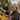 A joyful middle-aged Caucasian woman with blonde, wavy hair smiles warmly at the viewer, seen from an elevated perspective as she clears golden and brown autumn leaves from a dark house gutter. She wears a dark gray hooded sweatshirt and an orange-accented vest. Behind her, out of focus, a man with a beard operates a bright yellow gutter cleaning tool. The scene depicts a couple engaged in fall home maintenance on an overcast day, conveying a sense of collaboration and happiness.