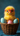 A fluffy, bright yellow baby chick wearing oversized bright pink eyeglasses is nestled in a brown wicker basket. It's surrounded by several colorful Easter eggs, including a pale pink egg held by its feet and two light blue eggs with tiny pink heart patterns. The background is a solid, deep teal color, creating a vibrant contrast.