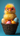 A bright yellow, extremely fluffy baby chick with tiny orange feet is centered in a dark brown wicker basket, prominently wearing oversized pink round eyeglasses. It is nestled among four decorated Easter eggs: one white with orange speckles, a pink one with white polka dots, a yellow one with white polka dots, and a light blue egg with subtle texture, all against a solid teal-blue background.