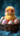 A fluffy yellow baby chick wearing bright pink-framed glasses sits prominently in a dark brown woven basket. It is surrounded by several speckled Easter eggs in soft pastel shades of pink, blue, and white, with orange accents. The background is a soft-focus, deep teal or blue fuzzy surface, creating a whimsical and adorable scene.
