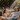 A father and his young daughter are joyfully laughing as they are splashed with water droplets under bright sunlight. They are sitting on an outdoor sofa, with the father reclined and the daughter sitting upright beside him, both wet and smiling widely. Lush green trees are blurred in the background, indicating an outdoor setting on a warm day.