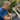 A middle-aged man with gray hair and a blue t-shirt is actively cleaning a white house gutter overflowing with dry brown and green autumn leaves. He wears gray work gloves and uses a tool to clear debris from the gutter, which runs along a dark shingled roof. Blurred green and yellow trees are visible in the background under natural daylight.