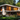 A modern two-story house with a white exterior, warm wooden slat siding, and large dark-framed windows stands prominently amidst a dense, lush green landscape. A flat roof with deep overhangs and a small balcony on the upper floor add to its contemporary design, while a stone path leads to the entrance. The house is surrounded by tall evergreen trees in the background and a variety of green shrubs and yellow flowers in the foreground, illuminated by bright daylight.