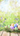 A vibrant vertical image featuring seven decorated Easter eggs in a natural bird's nest, positioned on light wooden planks. The eggs are pastel shades of green, purple, blue, and orange, each with white patterns. Lush green grass and delicate bluebell flowers surround the nest, with a soft, dreamlike bokeh background of greens and whites that creates a bright, cheerful spring scene.