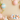 A pristine white rabbit with pink inner ears and nose sits calmly on a light beige surface, looking to the right. In front of it is a colorful string of knitted pom-poms, while a woven wicker basket filled with pastel blue, pink, and peach Easter eggs with delicate patterns sits to its right. Overhead, delicate branches with translucent pink, green, and yellow fabric leaves, along with horizontally striped pastel Easter eggs, frame the upper portion of the scene against the soft beige background.