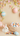 A pristine white rabbit with pink inner ears and nose sits calmly on a light beige surface, looking to the right. In front of it is a colorful string of knitted pom-poms, while a woven wicker basket filled with pastel blue, pink, and peach Easter eggs with delicate patterns sits to its right. Overhead, delicate branches with translucent pink, green, and yellow fabric leaves, along with horizontally striped pastel Easter eggs, frame the upper portion of the scene against the soft beige background.