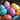 A close-up vertical shot of numerous brightly colored, speckled Easter eggs resting in a rustic twig nest. The eggs display a variety of hues including light pink, deep magenta, orange, yellow, and two shades of blue/teal, all dotted with white and smaller colored flecks. The background is softly blurred and dark, emphasizing the festive and cheerful eggs.