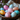 A close-up shot of several brightly colored Easter eggs in a dark, weathered wooden bowl. The eggs are decorated with various sized polka dots or speckles in pastel shades of pink, light blue, orange, and yellow. The bowl is resting on a textured beige linen fabric, with blurred purple flowers visible in the soft-focus background, suggesting a cozy and festive spring setting.