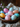 A close-up shot of several brightly colored Easter eggs in a dark, weathered wooden bowl. The eggs are decorated with various sized polka dots or speckles in pastel shades of pink, light blue, orange, and yellow. The bowl is resting on a textured beige linen fabric, with blurred purple flowers visible in the soft-focus background, suggesting a cozy and festive spring setting.
