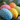 A close-up vertical image displays five brightly colored Easter eggs resting on green pine needles. The eggs are painted in a cheerful palette of blue, yellow, pink, orange, and green, each intricately decorated with numerous small white speckles. The foreground blue egg is in sharp focus, while the other eggs and the background are softly blurred, creating a warm and inviting spring holiday scene.