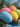 A close-up vertical image displays five brightly colored Easter eggs resting on green pine needles. The eggs are painted in a cheerful palette of blue, yellow, pink, orange, and green, each intricately decorated with numerous small white speckles. The foreground blue egg is in sharp focus, while the other eggs and the background are softly blurred, creating a warm and inviting spring holiday scene.