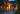 A family of three, a man, a woman, and a young boy, are seen from behind walking hand-in-hand towards a modern wooden house at dusk. The house is brightly lit with warm yellow lights, contrasting with the dark blue twilight sky and surrounding greenery. The path they walk on is gravel, lined with small rocks and subtle ground lighting, creating an inviting and peaceful scene of coming home.
