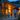 A family of three, consisting of a man, a woman, and a young boy, walks hand-in-hand away from the viewer on a gravel path towards a contemporary wooden house. The house is brightly lit with warm yellow light from inside and exterior fixtures, creating a welcoming glow against the dark blue twilight sky and distant green forest. Lush green foliage and decorative rocks line the pathway, with small ground lights also visible.