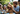 A heartwarming outdoor scene featuring a light brown, fluffy Goldendoodle dog wearing a pink polka-dot party hat, its tongue slightly out. A smiling man with curly hair and a beard gently pets the dog, while two young girls, also smiling, are partially visible on either side, one wearing a similar pink party hat. The background is a blurred green garden with subtle party banners, indicating a joyful family celebration around a table with a blue tablecloth.