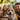 A close-up image features a happy tan and white dog wearing a festive party hat, looking directly at the viewer with an open-mouthed smile and pink tongue slightly out. Behind the dog, blurred figures of a smiling man and two young girls create a warm family scene outdoors, set against a soft, green bokeh background. The composition conveys a joyful and celebratory moment.