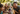 A close-up image features a happy tan and white dog wearing a festive party hat, looking directly at the viewer with an open-mouthed smile and pink tongue slightly out. Behind the dog, blurred figures of a smiling man and two young girls create a warm family scene outdoors, set against a soft, green bokeh background. The composition conveys a joyful and celebratory moment.