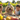 A fluffy golden-cream dog wearing a colorful birthday hat sits on a wooden table, surrounded by a smiling young girl, boy, and adult man. The outdoor scene is bathed in warm golden light, conveying a happy family birthday celebration for their pet.