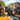 A joyful outdoor birthday party scene featuring a diverse family and their pet. A bearded man, two young girls, a young boy, and a shaggy black dog, all wearing colorful party hats, laugh heartily around a table with blurred party food in the foreground. Bright sunshine illuminates their happy faces against a backdrop of green trees and festive bunting, creating a warm and celebratory atmosphere.
