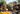 A joyful outdoor birthday party scene featuring a diverse family and their pet. A bearded man, two young girls, a young boy, and a shaggy black dog, all wearing colorful party hats, laugh heartily around a table with blurred party food in the foreground. Bright sunshine illuminates their happy faces against a backdrop of green trees and festive bunting, creating a warm and celebratory atmosphere.