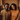 A medium shot of a happy young Black couple smiling brightly at the camera. They are standing close together in a room filled with cardboard moving boxes, suggesting they have just moved into a new home. The man is on the left, wearing a textured tan t-shirt, and the woman on the right, wearing a textured orange sweater and hoop earrings.