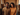 A medium shot of a happy young Black couple smiling brightly at the camera. They are standing close together in a room filled with cardboard moving boxes, suggesting they have just moved into a new home. The man is on the left, wearing a textured tan t-shirt, and the woman on the right, wearing a textured orange sweater and hoop earrings.